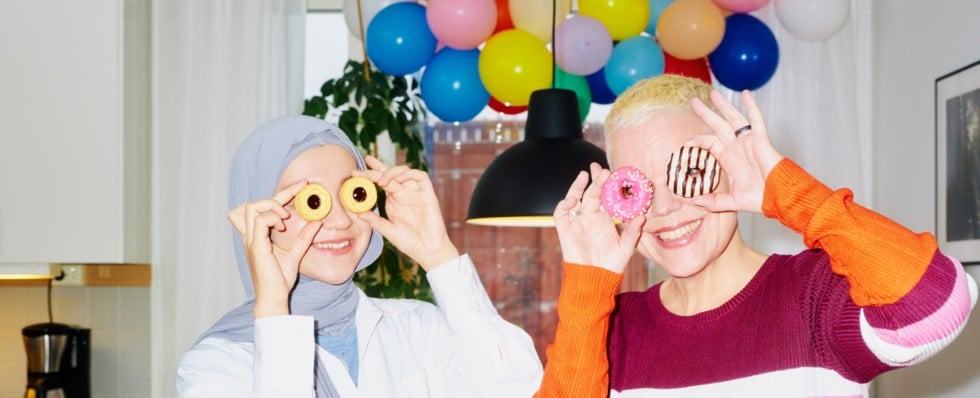 Två tjejer står framför en färgglad bakgrund av ballonger. Den ena personen bär en grå hijab och vit skjorta och håller kakor framför sina ögon. Den andra personen har kort blont hår och en randig tröja i orange, rosa, vit och lila och håller munkar framför sina ögon. Båda ler.